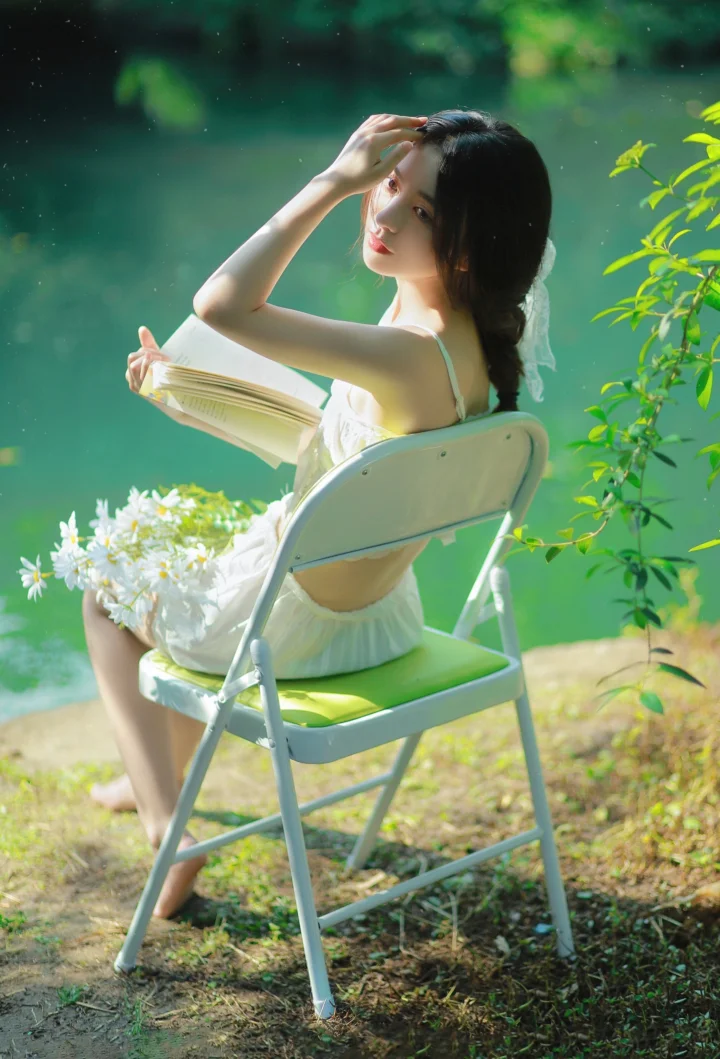 한 여성이 호숫가 접이식 의자에 앉아 책을 들고 흰 꽃과 푸른 잎사귀에 둘러싸여 있다.
