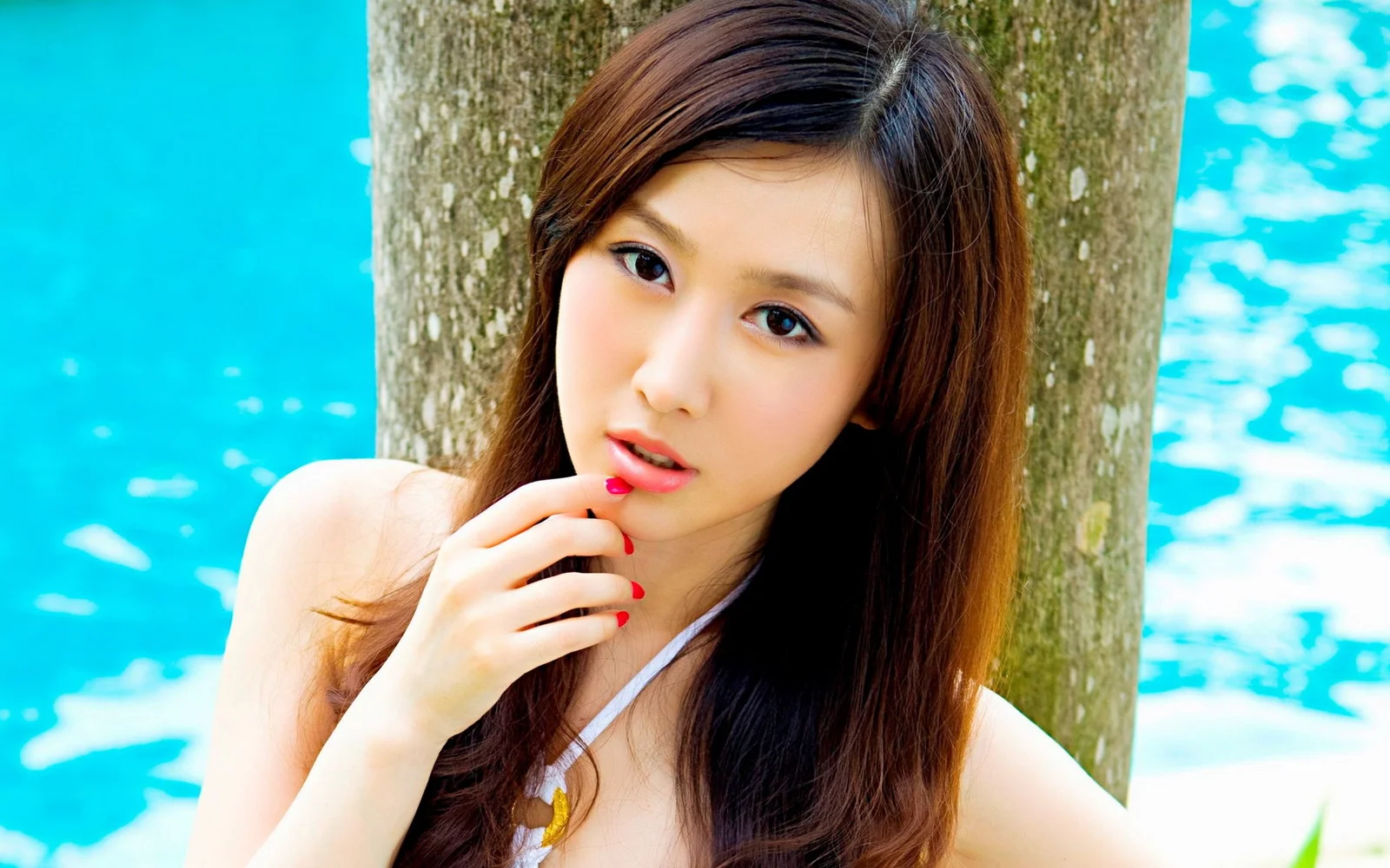一位成年女性站在树旁，身后是明亮的蓝色水面。