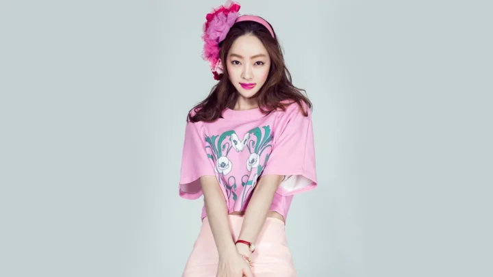 분홍색 상의와 꽃 장식을 착용한 성인 여성이 연한 회색 배경 앞에서 포즈를 취하고 있습니다.