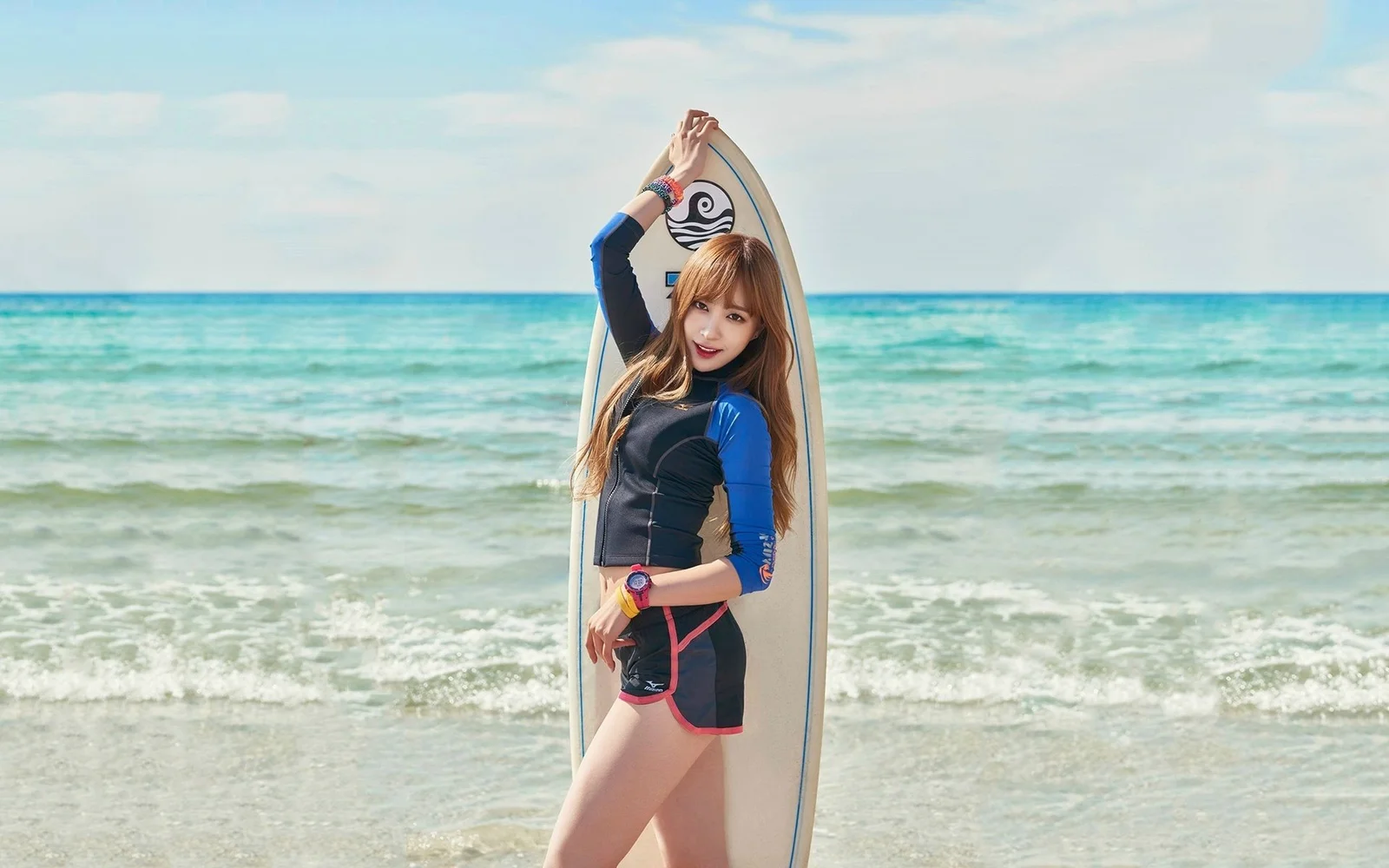 大人の女性がサーフボードを持って海辺に立ち、背後に明るい海と砂浜が広がっています。