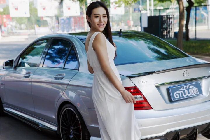 一名身穿白色长裙的成年女子站在户外银色豪华轿车旁。