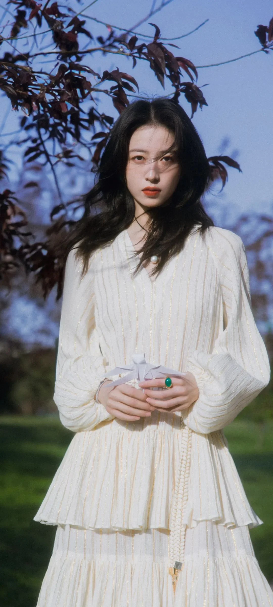긴 머리의 여성이 흰색 긴소매 원피스를 입고 나무와 잔디를 배경으로 야외에 서 있다.