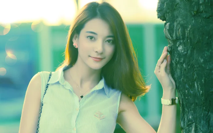 一位长发女子站在树旁，身穿浅色无袖衬衫，沐浴在柔和的绿色户外光线中。