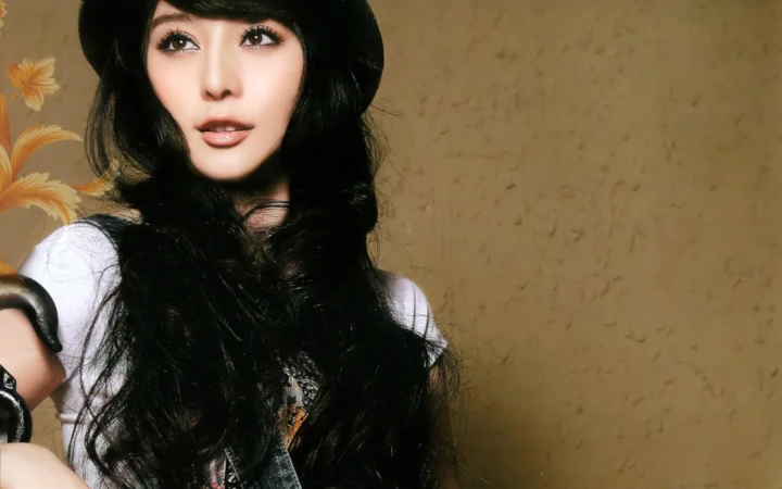 一名黑发女性面对镜头，背景是棕色墙面和左侧的花卉图案。