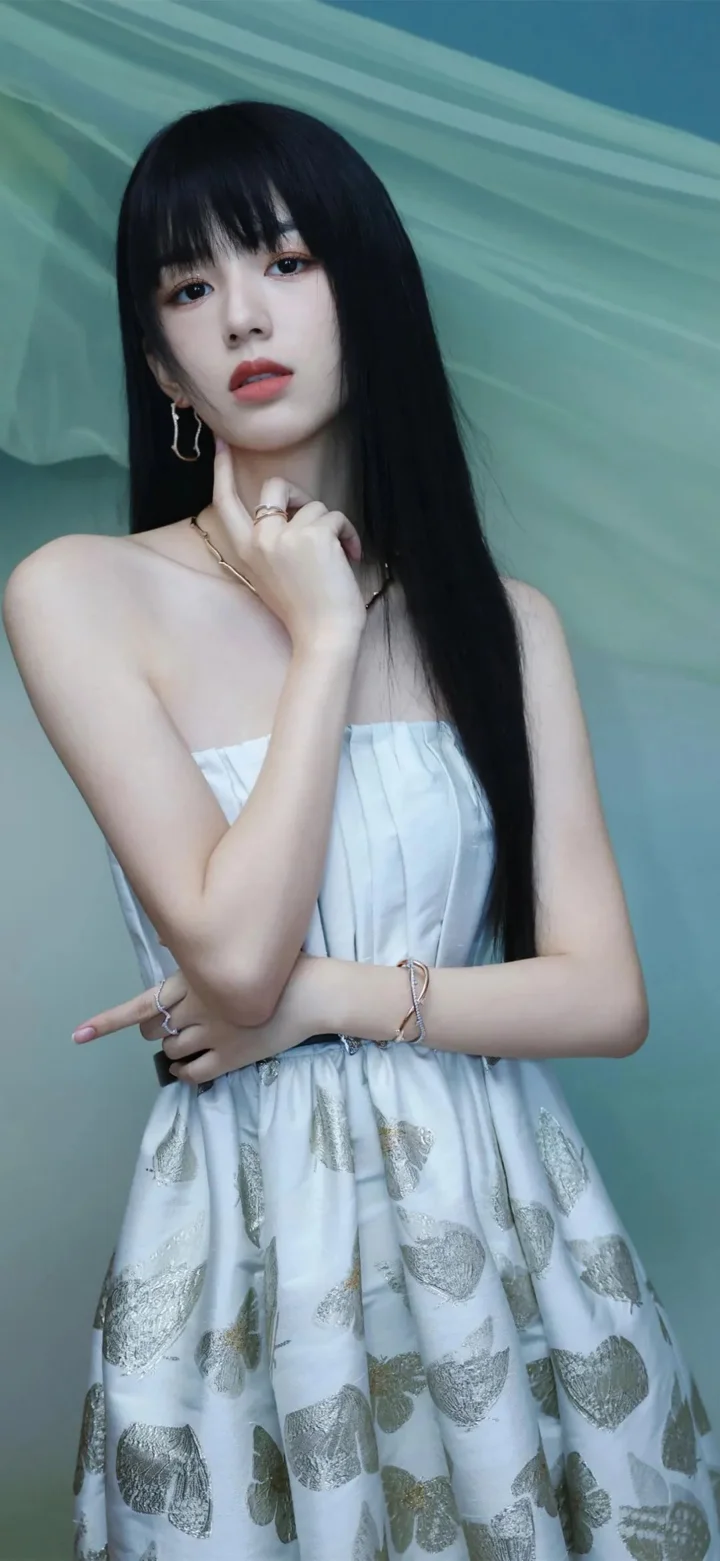 검은 긴 머리의 여성이 흰색 민소매 드레스를 입고 연한 청록색 배경 앞에서 포즈를 취하고 있다.