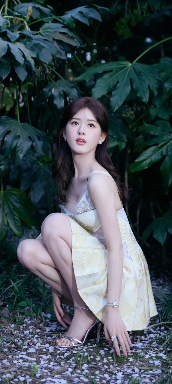 一名成年女性穿着白色连衣裙，蹲坐在茂密绿叶和散落花瓣之间。