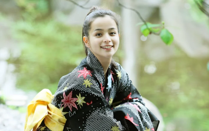 一名身穿黑色花纹和服的女子坐在绿色森林中微笑面对镜头。