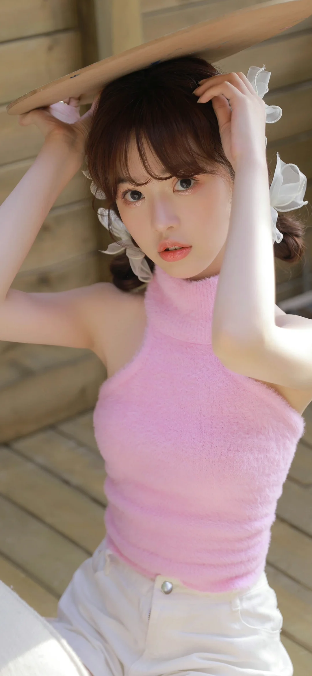 一名成年女性身穿粉色无袖上衣，在木质背景前抬手整理头发。