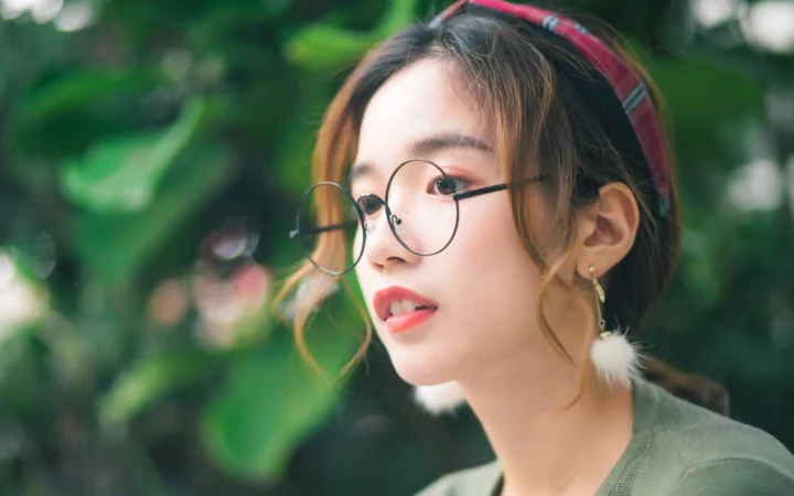 一位戴圆框眼镜的女性在绿叶背景前侧脸望向一旁。