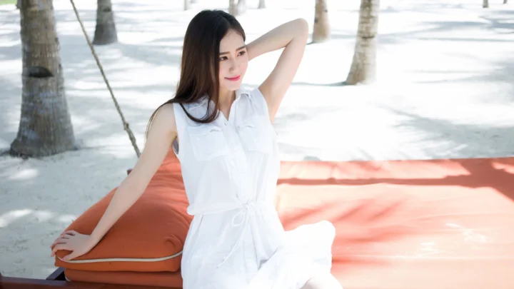 一名身穿白色连衣裙的女子坐在户外橙色垫子上，背景是白色沙地和棕榈树。