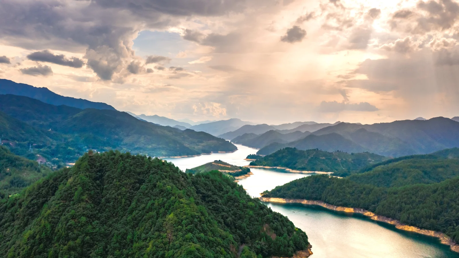 雲間から差し込む夕日が、連なる山々と森林、曲がりくねった湖を照らしています。