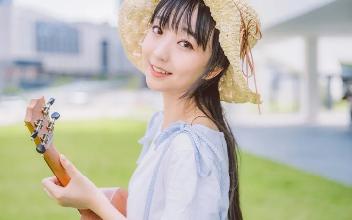 一名戴草帽的女性在户外草地旁手持尤克里里微笑面对镜头。