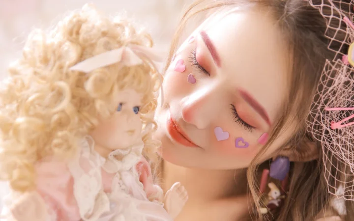 눈을 감은 여성이 금발 인형에 가까이 기대고 있으며 얼굴에는 분홍색 하트 스티커가 붙어 있다.