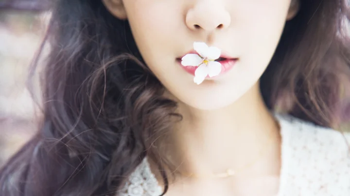 긴 머리 여성이 입술에 작은 흰 꽃을 문 클로즈업 인물 사진입니다.