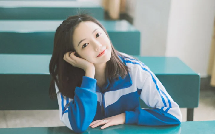 一名长发女性身穿蓝白运动服，坐在教室座位旁托腮看向镜头。