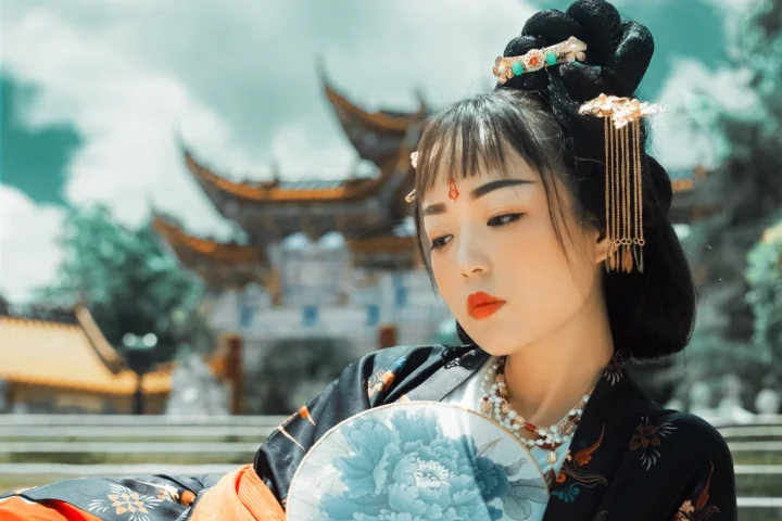 一位身穿传统服饰的成年女子手持牡丹图案折扇，站在古建筑前。