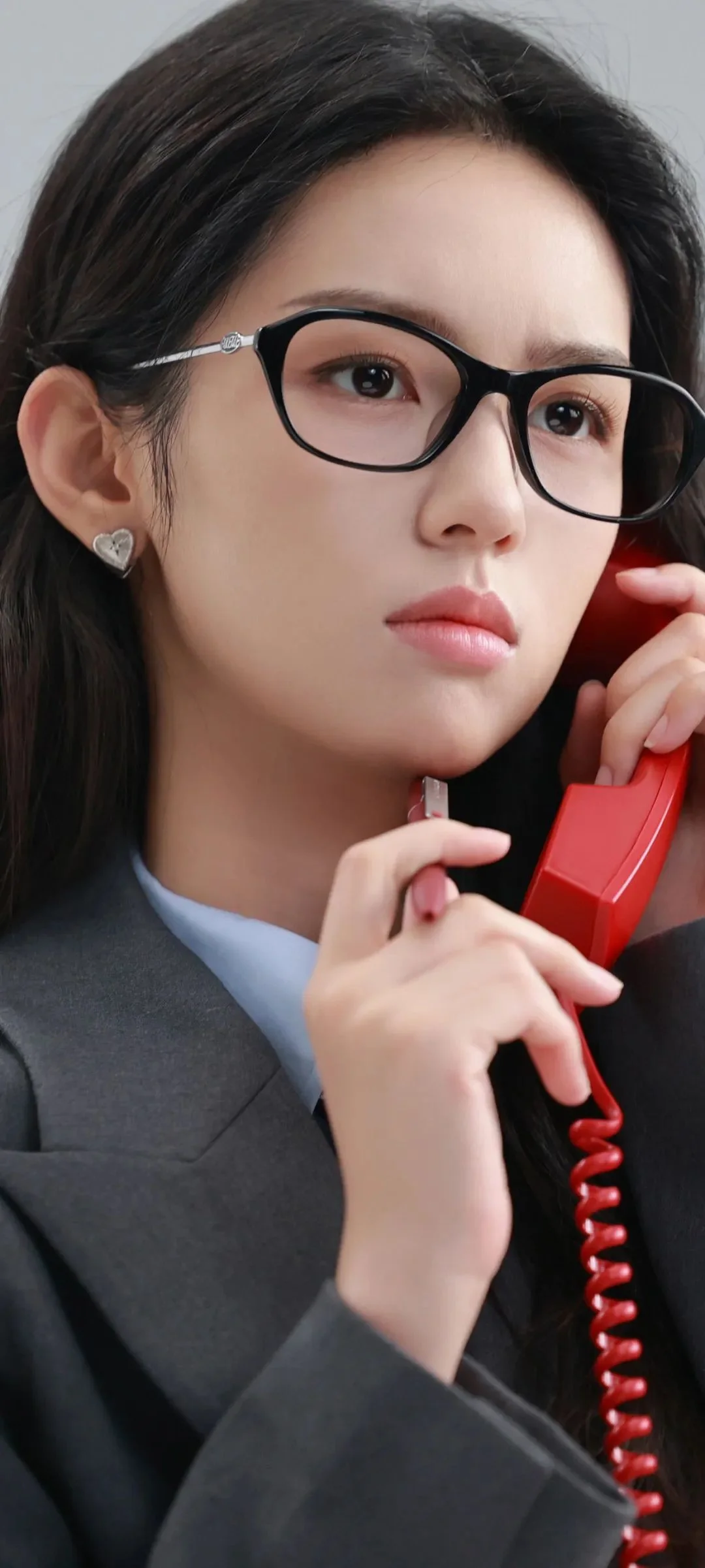 一名戴黑框眼镜的女子手持红色电话听筒，身穿深色服装，画面为竖幅近距离肖像。