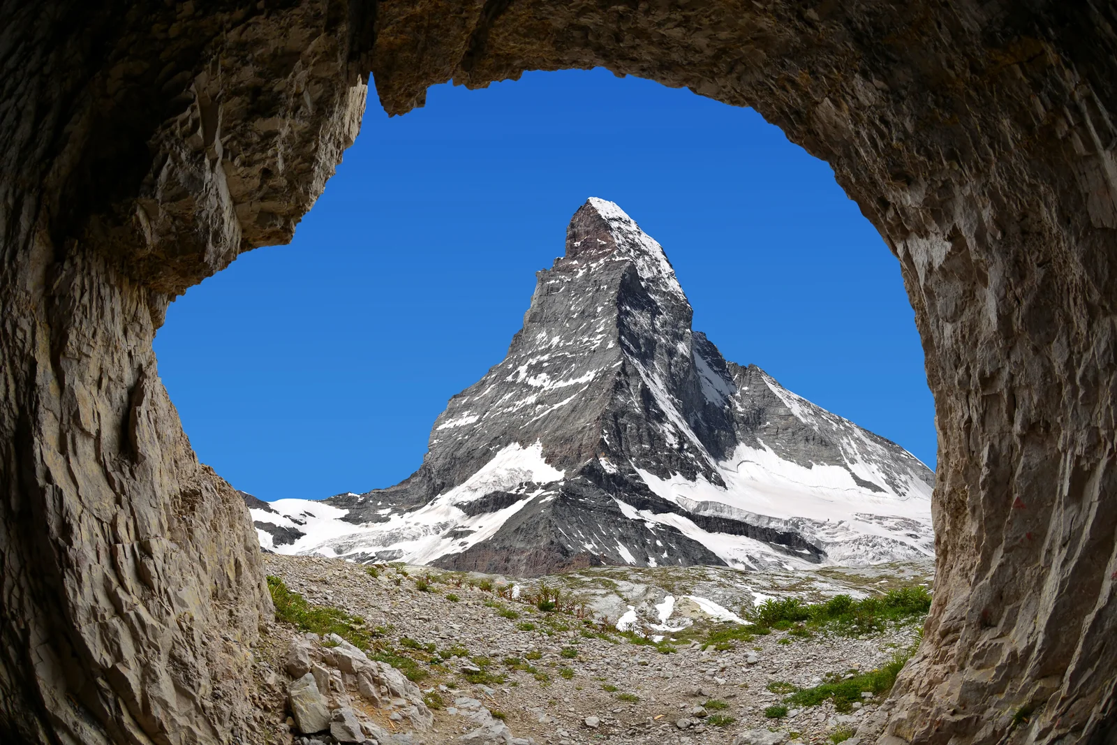 从岩洞中望去，一座积雪覆盖的尖峭山峰矗立在晴朗的蓝天下。