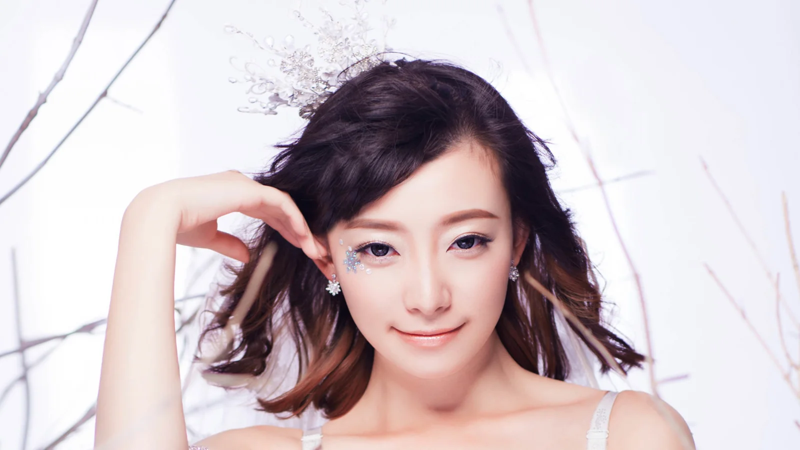 밝은 흰색 배경 앞에서 섬세한 메이크업과 짙은 웨이브 머리를 한 성인 여성이 포즈를 취하고 있습니다.