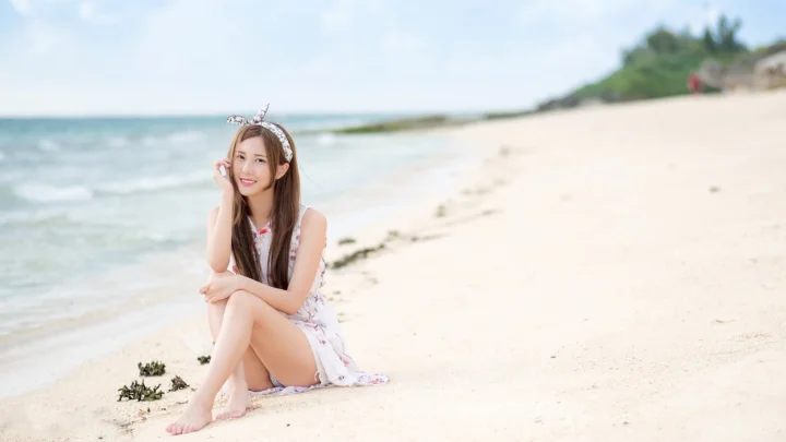一名成年女性坐在海滩上，身后是海水、沙滩和明亮的天空。
