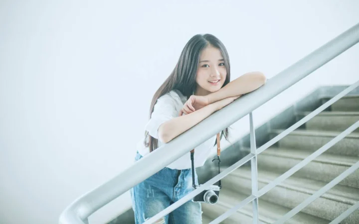 一名微笑的女性倚靠在楼梯扶手上，身旁悬挂着一台相机。