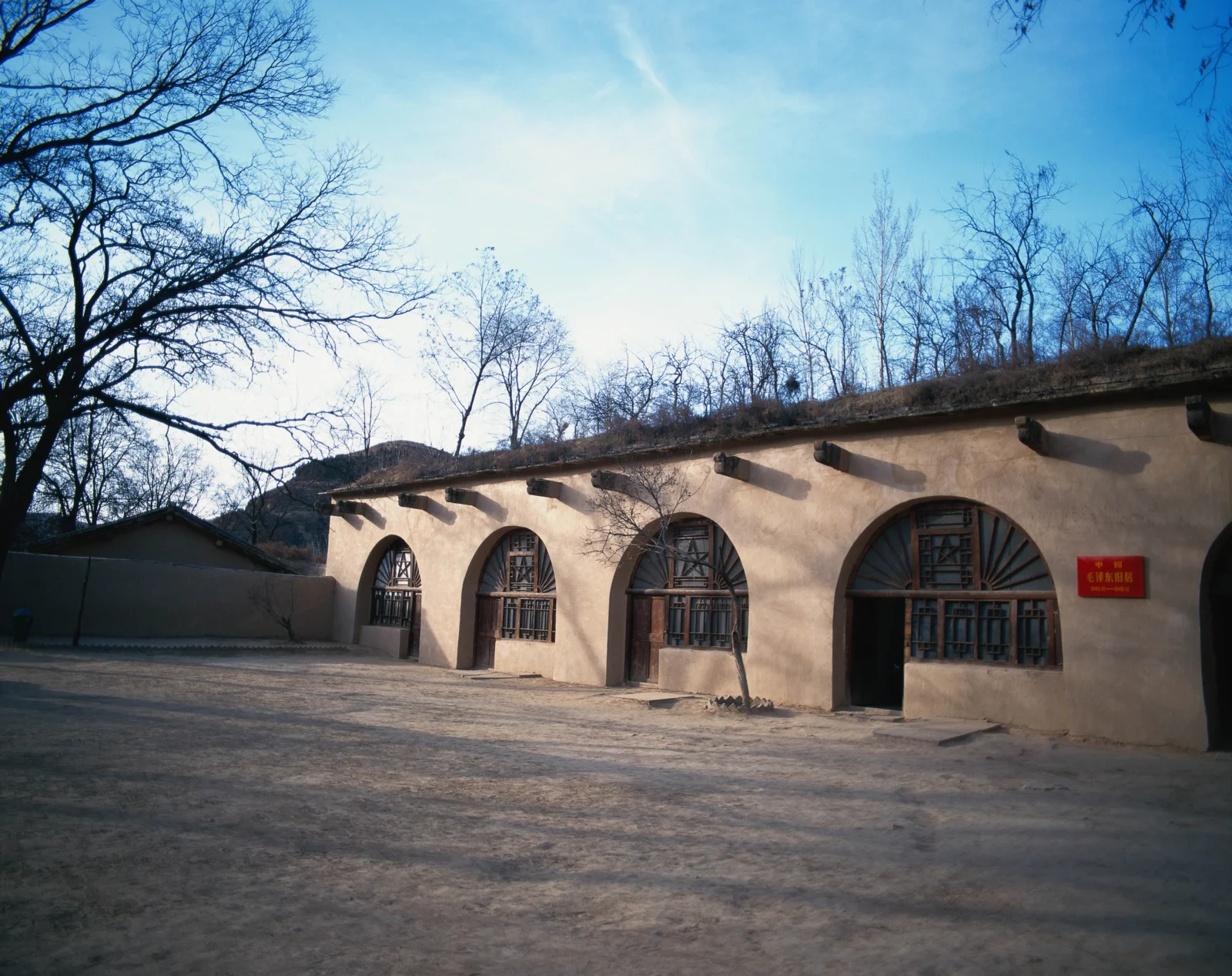 一座黄土色古建筑立于铺装庭院旁，墙面设有拱形窗户，周围可见冬季的无叶树木和蓝天。