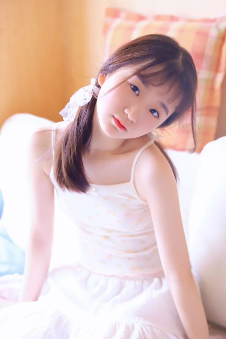 一位棕色长发女性身穿白色无袖连衣裙，坐在床上接受柔和光线照射。
