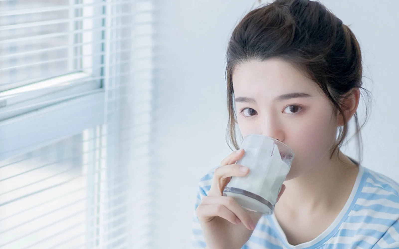 一名女子站在窗边，手持玻璃杯饮水。
