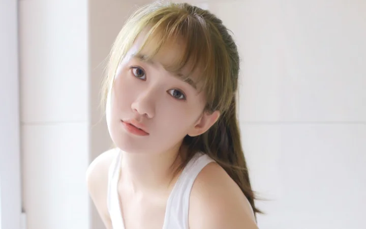 갈색 머리 여성이 흰색 상의를 입고 밝고 단순한 배경 앞에서 카메라를 향해 고개를 살짝 기울이고 있습니다.