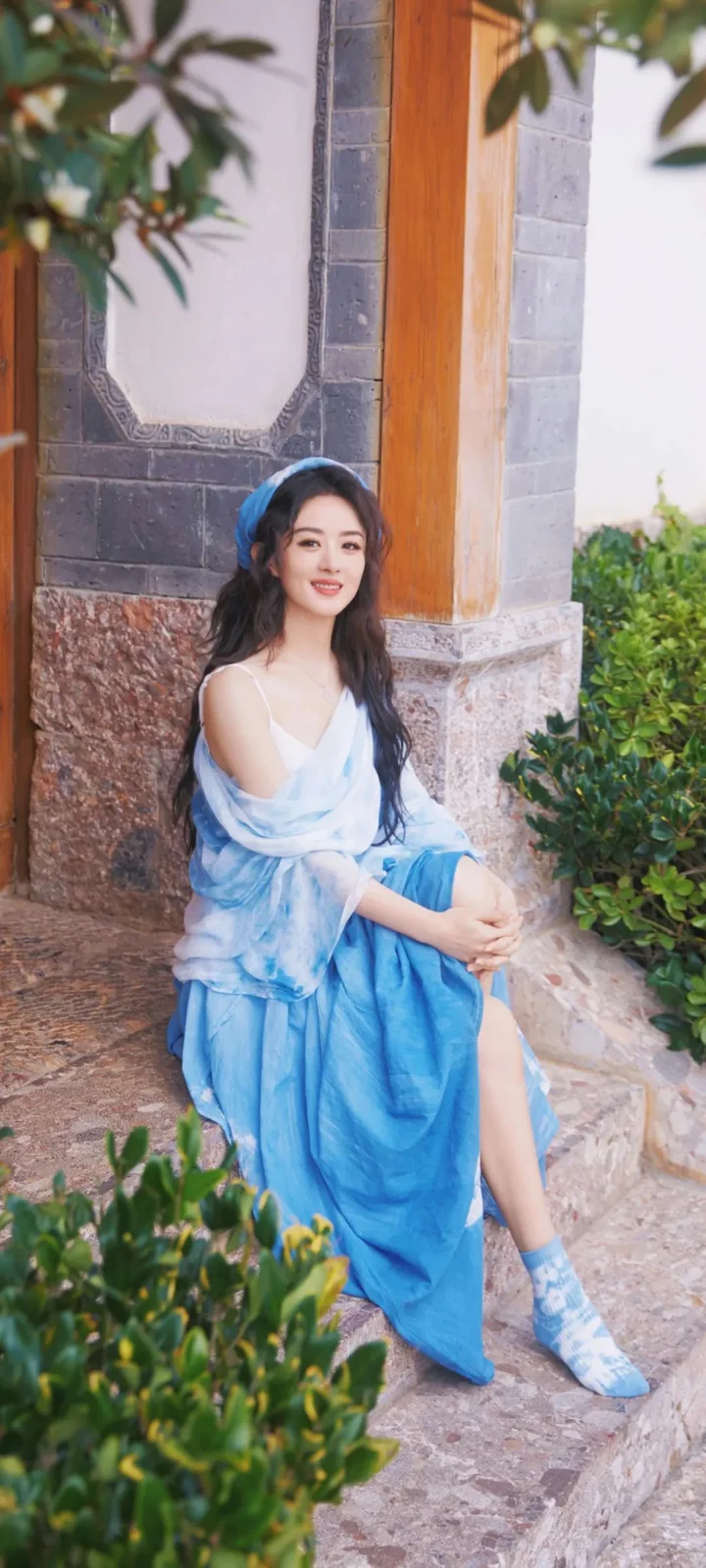 一名身穿蓝色长裙的女子坐在石墙与木门旁，周围有绿植环绕。