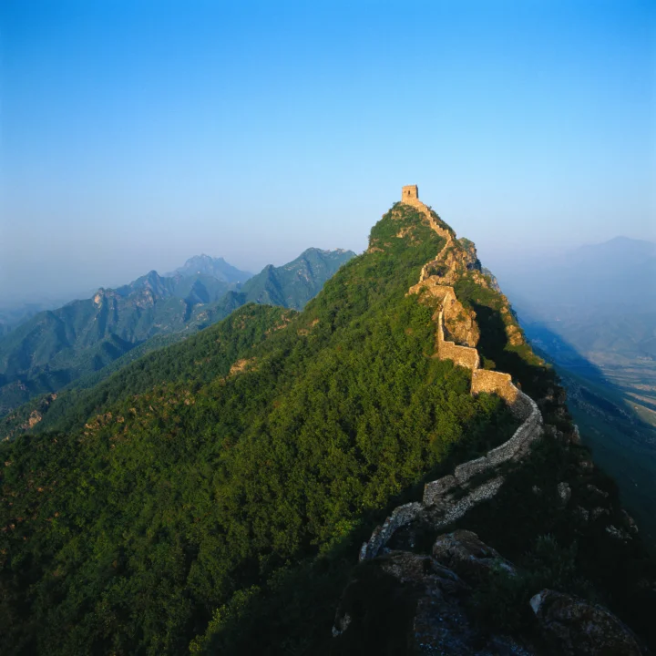 蜿蜒的长城沿着覆满森林的山脊延伸，远处是层叠群山与晴朗蓝天。