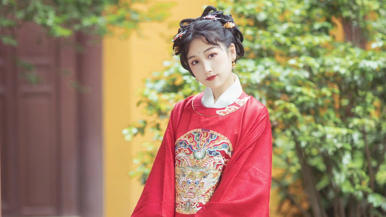 정교한 자수가 있는 붉은 전통풍 의상을 입은 여성이 초목으로 둘러싸인 안뜰에 서 있습니다.