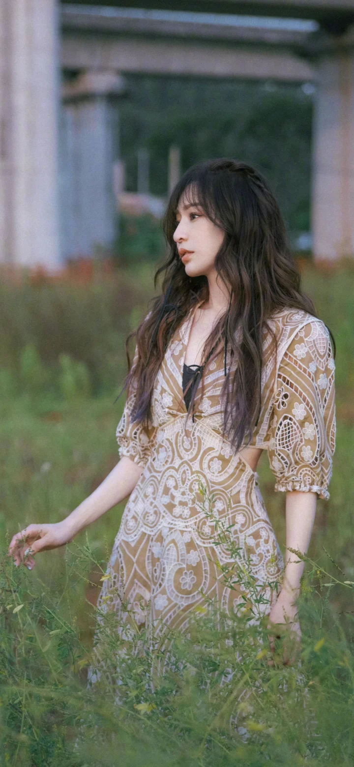 一名长发女子穿着图案连衣裙站在草地上，身后是混凝土结构和树木。