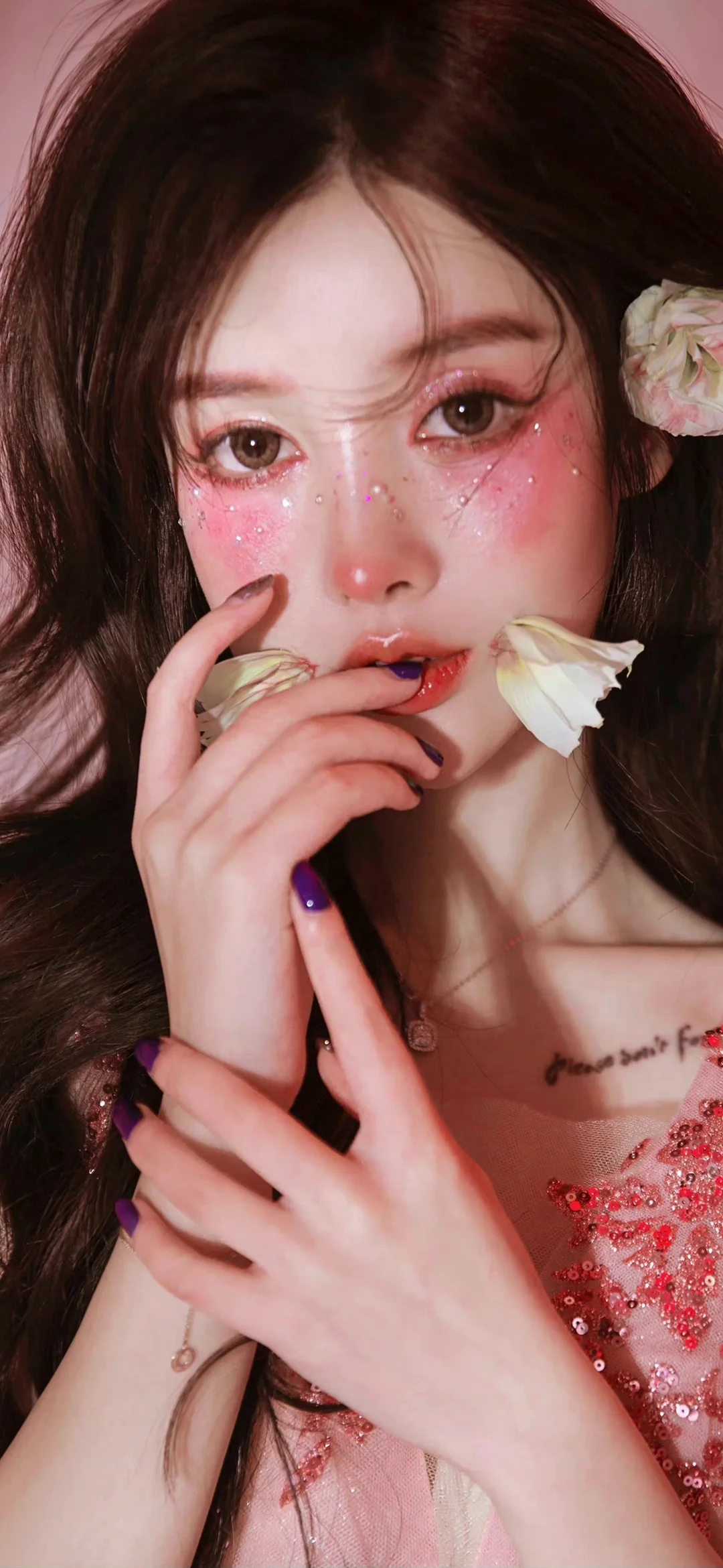 한 여성이 카메라를 가까이 바라보고 있으며, 볼에는 분홍색 메이크업과 반짝이가 있고 머리카락과 입가에 흰 꽃이 장식되어 있습니다.