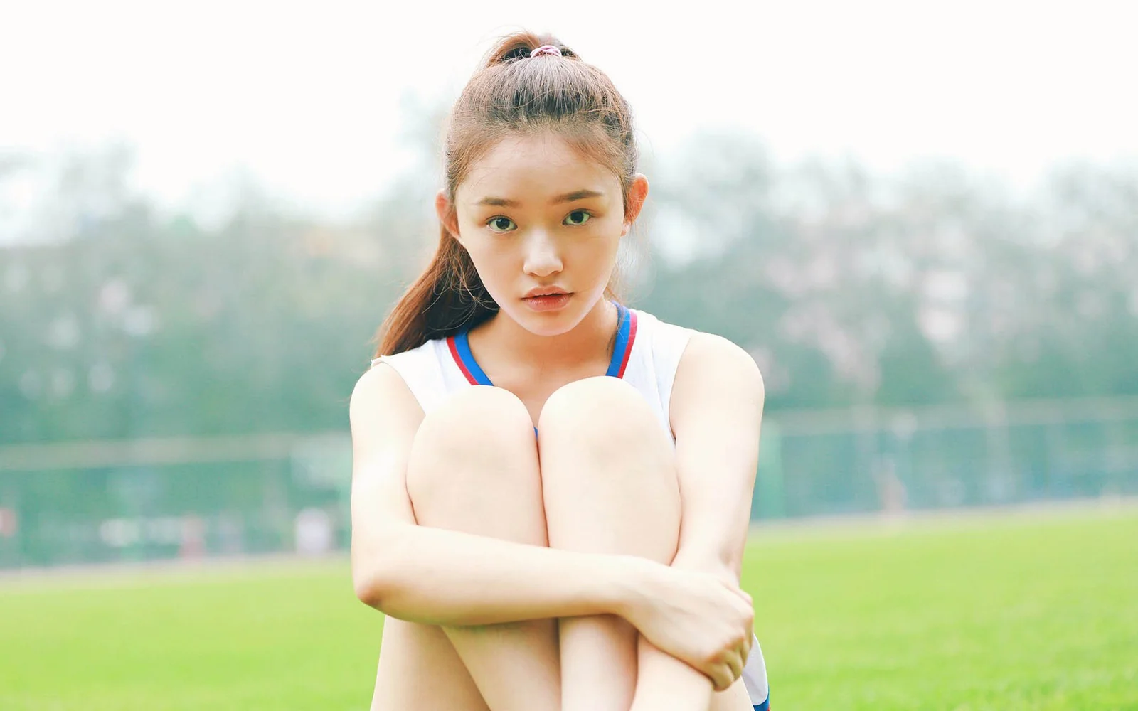한 여성이 잔디밭에서 무릎을抱고 앉아 있으며 뒤로 흐릿한 나무와 수면이 보입니다.
