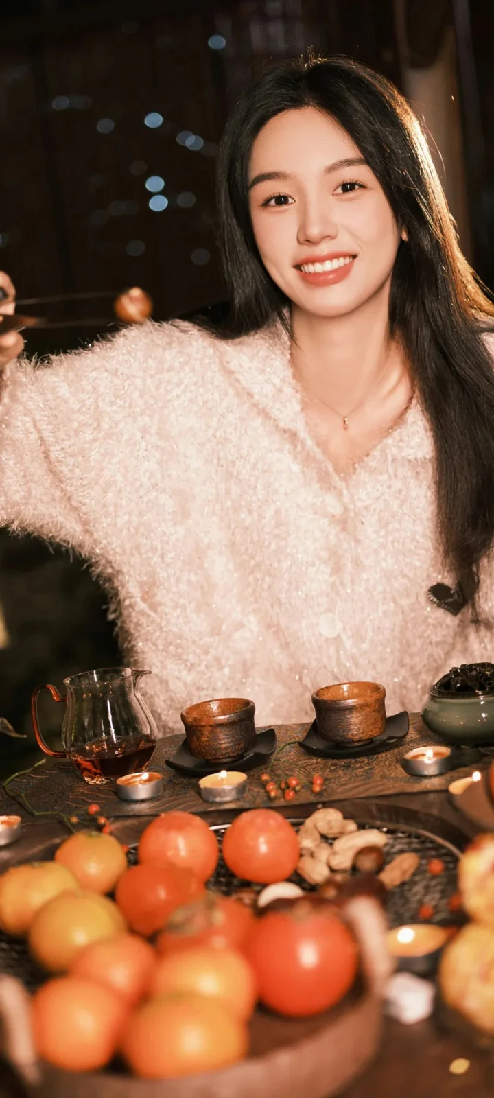 긴 머리의 여성이 밝은 색 스웨터를 입고 차 도구와 과일이 놓인 실내 테이블 옆에서 미소 짓고 있다.
