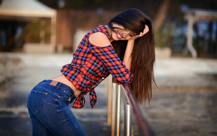 一名身穿红色格纹衬衫和牛仔裤的女性在户外俯身微笑。