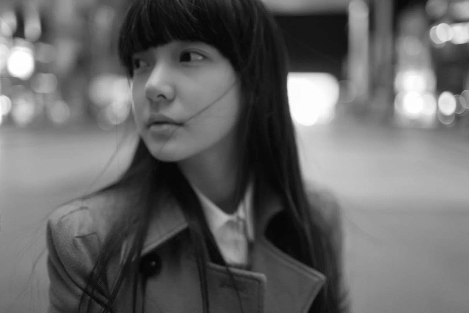 일자 앞머리의 성인 여성이 거리에 서 있으며 흑백 배경의 불빛이 부드러운 보케로 표현되어 있다.