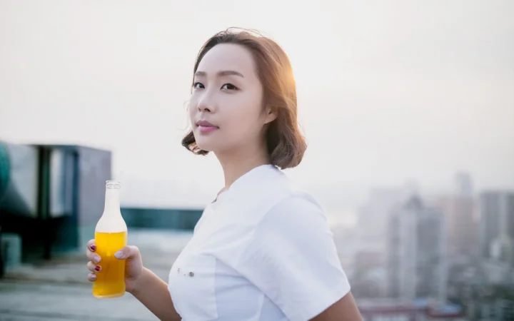 흰색 상의를 입은 성인 여성이 노란 음료가 담긴 병을 들고 있으며 뒤로 흐릿한 도시 풍경이 보입니다.