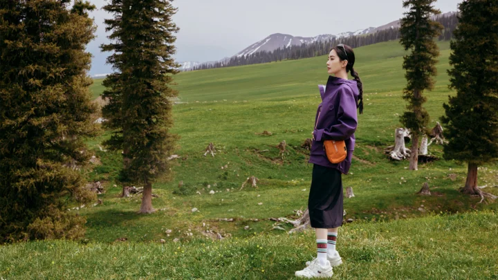 一名身穿紫色外套的女子站在绿意盎然的山间草甸上，远处是积雪山脉和高大树木。