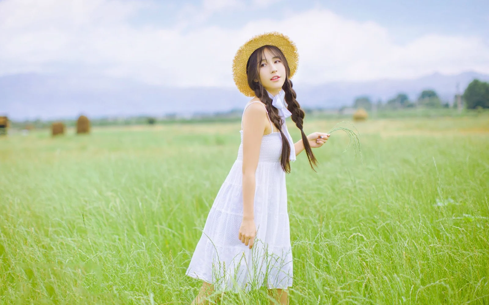 一名戴著草帽、穿著白裙並留著長辮子的女子站在綠色草地中。