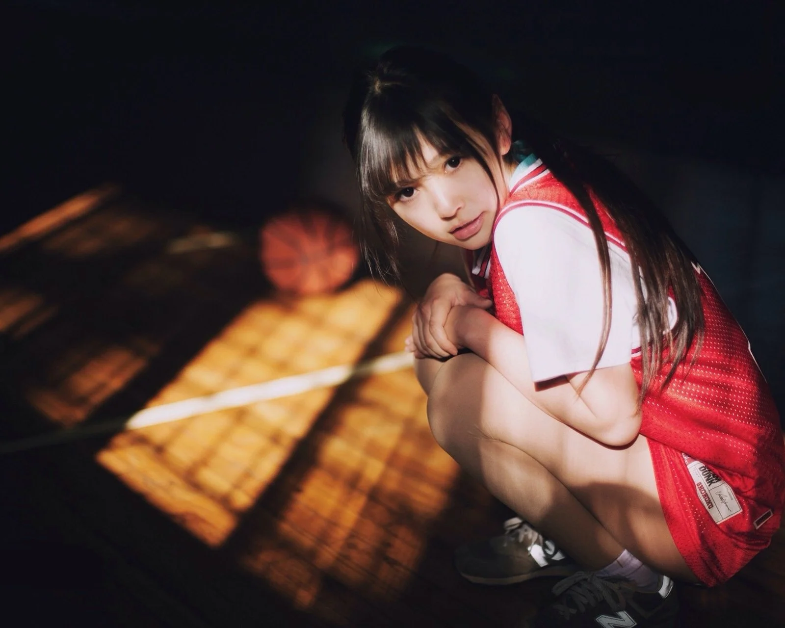 赤いスポーツウェアを着た女性が、床に暖かな光と影が映る暗い場所でしゃがんでいる。