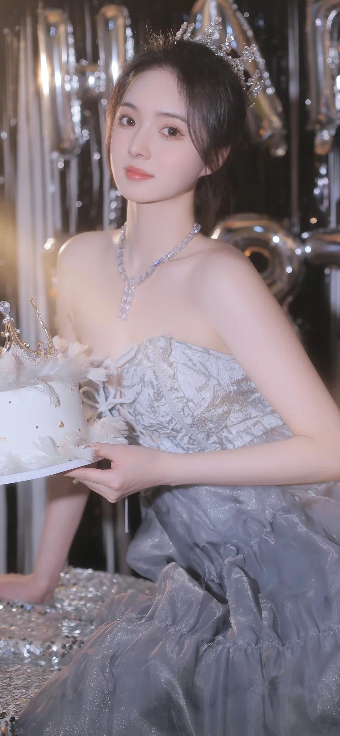 銀灰色のドレスを着た女性が、きらめく装飾の前でケーキを持ってポーズを取っている。