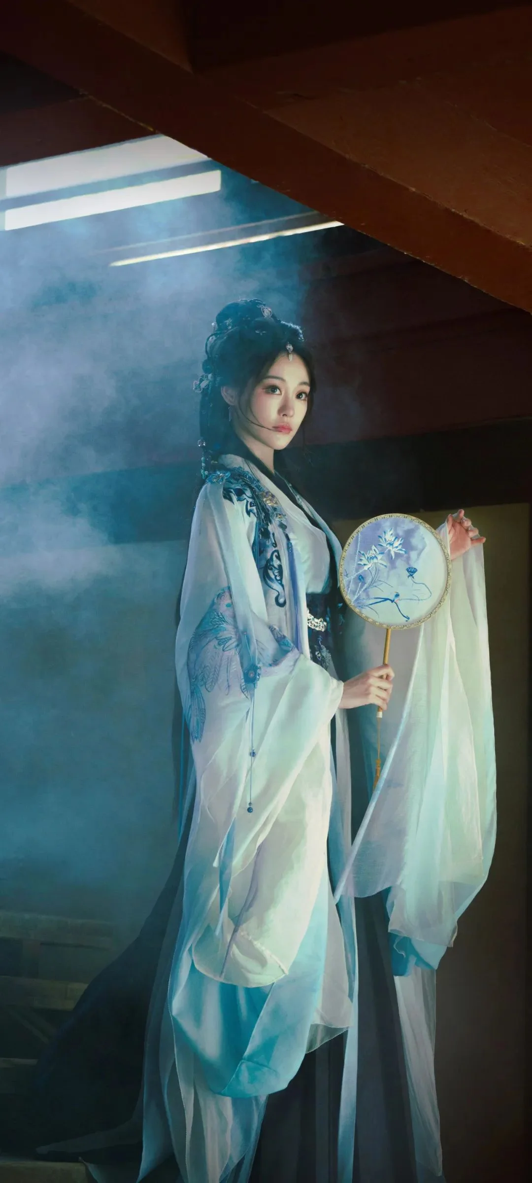 一位身穿浅色传统服饰的女子在烟雾缭绕的室内手持团扇。
