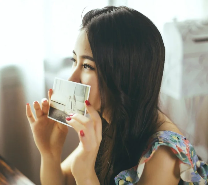 一名长发女性在窗边手持一张照片靠近脸部。