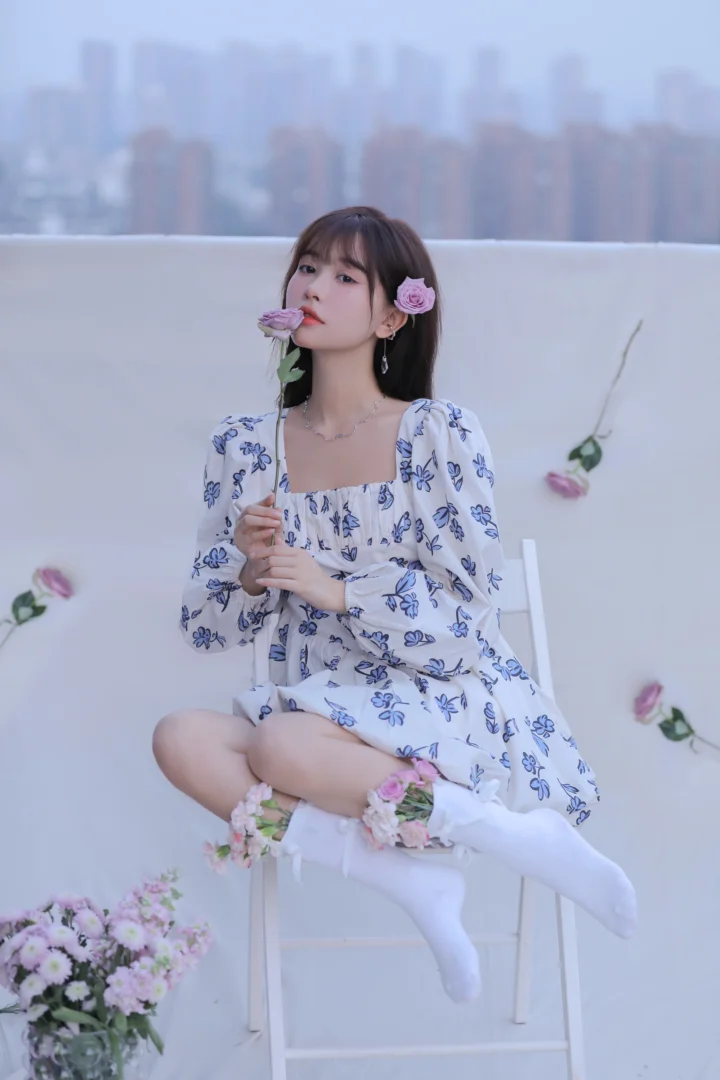 一名成年女子坐在白色椅子上，身穿白色花卉洋裝和白色長襪，手持一朵玫瑰，背景是模糊的城市天際線。