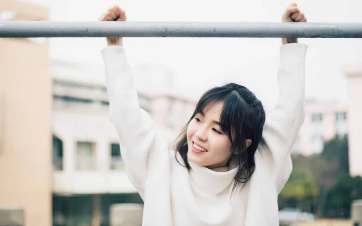 흰색 스웨터를 입은 여성이 야외에서 가로봉을 잡고 있으며 배경에는 흐릿한 건물이 보입니다.