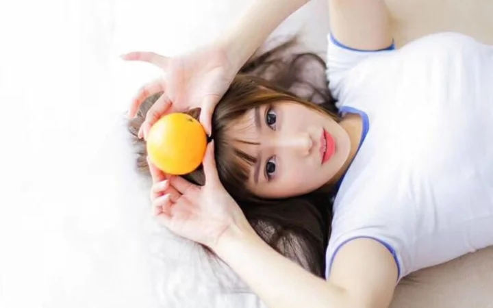 흰색 상의를 입은 젊은 여성이 흰 표면에 누워 머리 위로 오렌지를 들고 있다.