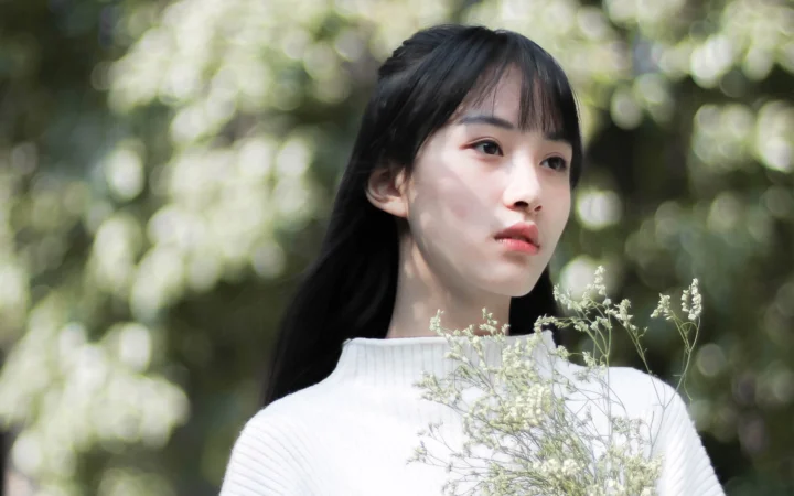 一名黑发女子身穿白色上衣站在林间，身前映入几枝细小的白色野花。