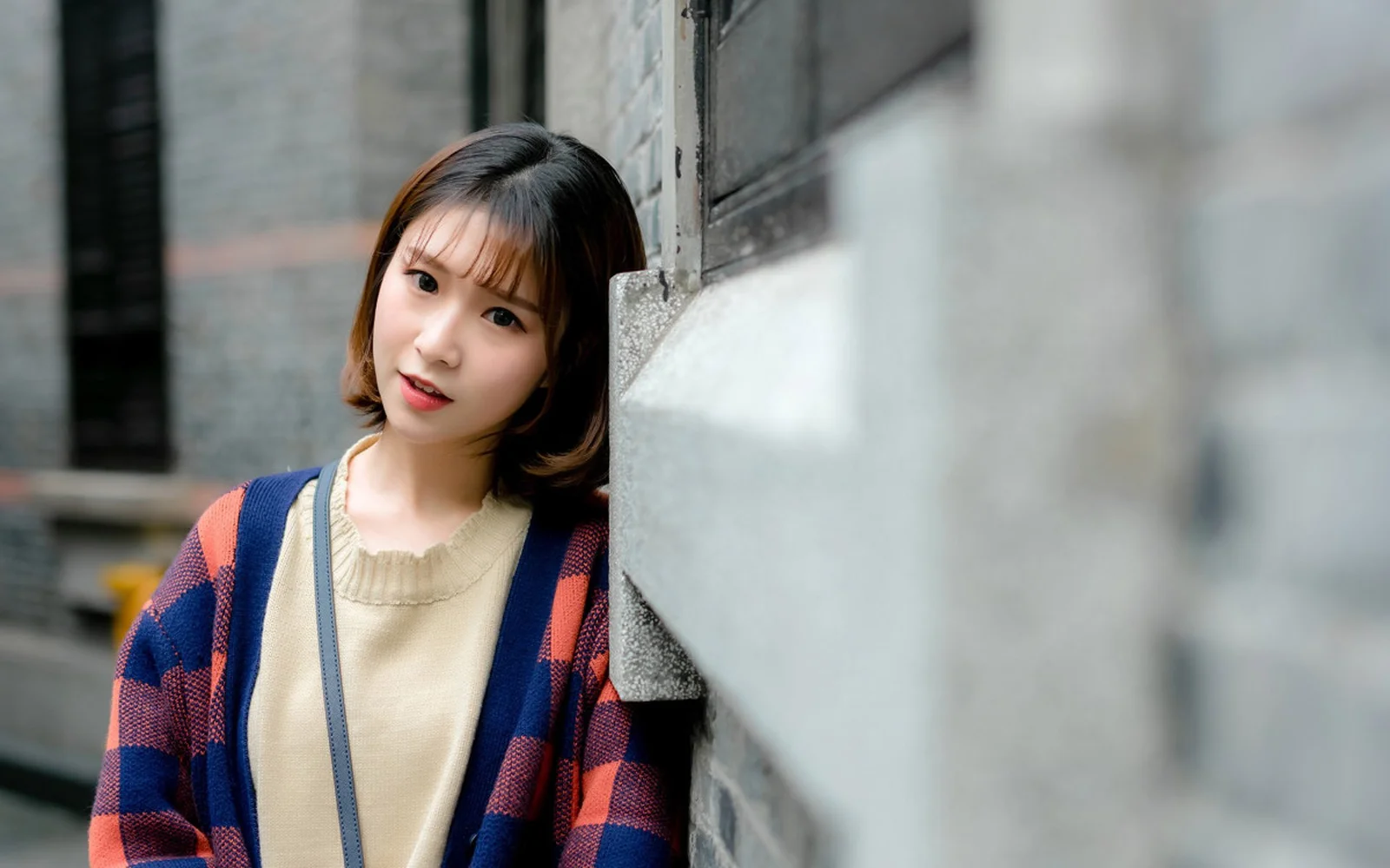 단발머리 여성이 도시 건물 벽 옆에 서서 카메라 쪽으로 살짝 몸을 향하고 있다.